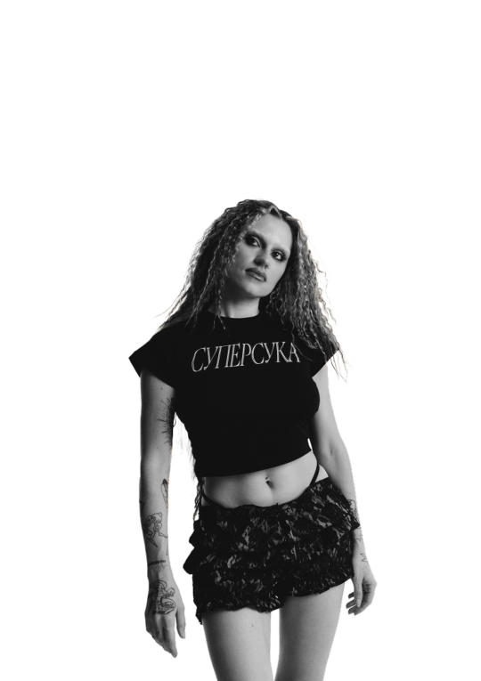 Portrait of a woman with curly hair wearing a black cropped shirt and lace shorts, standing against a solid black background (shirt reads Cyrillic text).